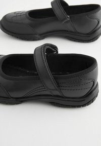 Chaussures en cuir noir avec une sangle en Velcro, bout rond et semelle en caoutchouc. Présentent des coutures visibles et un design texturé sur la partie supérieure.