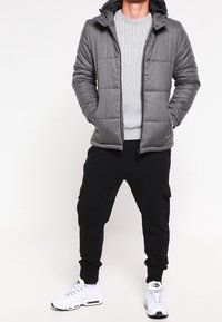 Grå quiltet dunjakke med sort hætte, båret over en lys grå kabelstrikket sweater, kombineret med sorte cargo-joggers og hvide sneakers.