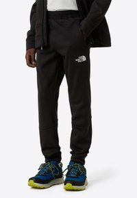 Svarte joggebukser laget av mykt stoff, med en logo på låret, elastisk midjebånd og smale ben. Kombinert med blå og svarte sko.