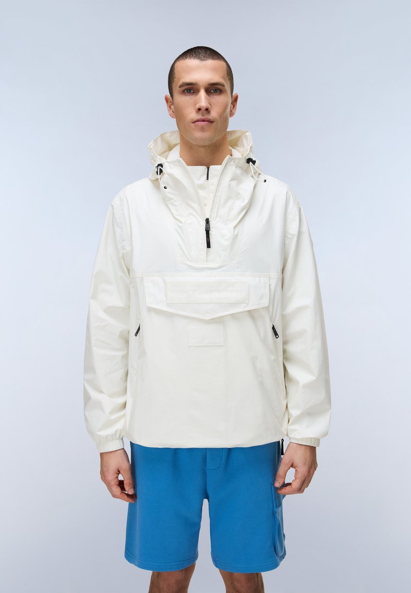 Άνδρας που φοράει λευκό αδιάβροχο μπουφάν με κουκούλα και μπροστινή τσέπη, καθώς και μπλε σορτς, στέκεται μπροστά σε ένα απλό φωτεινό φόντο.