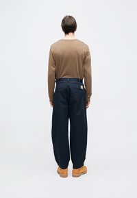 Brązowa koszula z długim rękawem i okrągłym dekoltem, zestawiona z luźnymi, ciemnoniebieskimi spodniami. Charakterystyczna pomarańczowa metka na tylnej kieszeni.