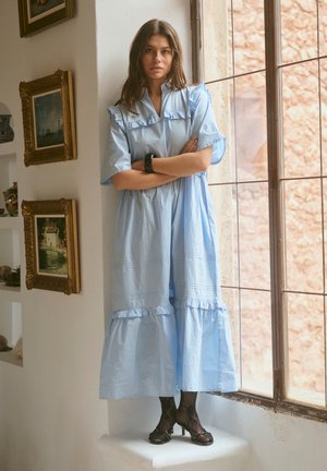 Világoskék fodros ruhát viselő nő fekete csipkenadrágban és magassarkúban, karját összefonva, egy ablak mellett áll, mögötte a falon keretezett festmények láthatók.