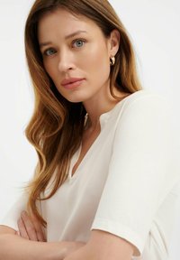 Witte V-hals top met korte mouwen, gemaakt van zacht materiaal. Model heeft steil bruin haar en draagt gouden ringoorbellen. Neutrale achtergrond.