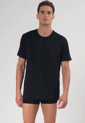 Jonge man die staat te poseren in een eenvoudig zwart t-shirt met korte mouwen en zwarte shorts tegen een lichtgrijze achtergrond.