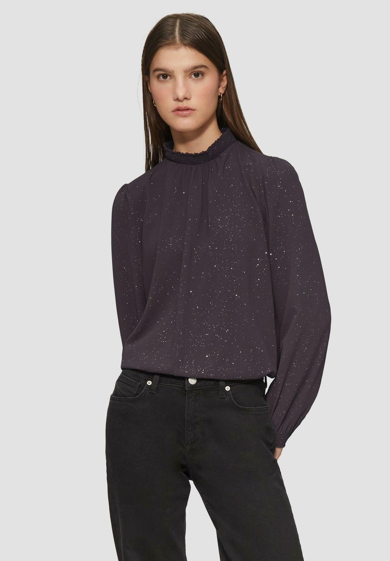 Dunkellila Bluse mit hohem Rüschkragen, langen Ärmeln und subtilen Sprenkelmuster. Leichter Stoff mit glatter Textur.
