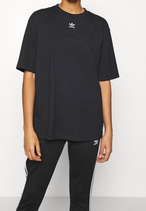 Persona que lleva una camiseta negra oversize de Adidas y pantalones deportivos negros Adidas con rayas blancas, de pie frente a un fondo liso y claro.