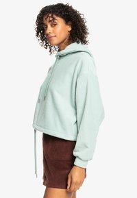 Sudadera corta verde menta con cordones y una textura suave, combinada con una falda de pana marrón con bolsillos frontales.