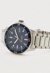 Roostevabast terasest käekell sinise ja kuldse äärega, tumeda sinise hõlma, kollaste tunni indikaatoritega, kuupäevaaken kl 3 ja metallist lingirihmaga.