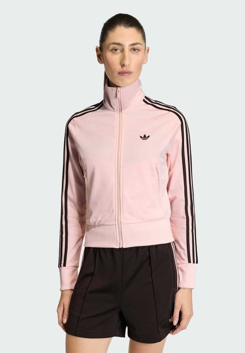Ružová tréningová bunda so zipsom, čierne pruhy na rukávoch a malým čiernym logom na hrudi. Vyrobená z hladkej, ľahkej látky.