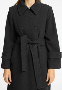 Črni plašč v ribji kost z dvojnim zapenjanjem, velikim ovratnikom in pasom. Ima gumbne manšete in teksturirano tkanino.