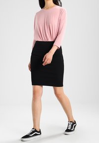 Nainen, jolla on päällään vaaleanpunainen pitkähiha blouse, musta polviin ulottuva hame ja mustat tennarit valkoisilla nauhoilla, kävelee yksinkertaista valkoista taustaa vasten.