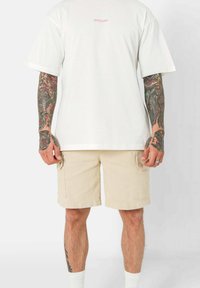 T-shirt bianca in cotone con maniche corte, che presenta un testo del logo rosa. Abbinata a pantaloni cargo beige, con tasche e texture in cotone.