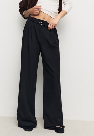 Person trägt schwarze, hoch taillierte, weit geschnittene Hose mit Gürtel, weißes, kurz geschnittenes Langarmshirt und schwarze Ballerinas, steht vor einer schlichten weißen Wand.