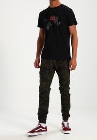 Černé tričko s lesklou grafikou v červené a šedé barvě, spárované s maskáčovými joggers a červenými teniskami Vans. Hladký materiál.