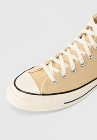 Μπεζ υφασμάτινο αθλητικό παπούτσι ψηλού τύπου με λευκά κορδόνια, καουτσούκ κάλυμμα δακτύλων και μαύρη ρίγα διακόσμησης. Διαθέτει  μεταλλικά ρούμπελα και υφή στη σόλα.