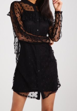 Kobieta w czarnej sukience zapinanej na guziki z przezroczystymi długimi rękawami w groszki i falbankami, pozująca z jedną ręką przy twarzy.