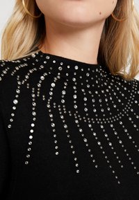 Schwarzer Pullover mit hohem Kragen, verziert mit silbernen Strasssteinen, die in einem kaskadierenden Muster angeordnet sind, und eine strukturierte Oberfläche aufweisen.