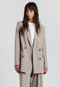 Donna con lunghi capelli scuri indossa un blazer beige doppiopetto e pantaloni abbinati su una maglietta bianca, in piedi contro uno sfondo chiaro e semplice.