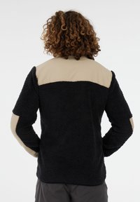 Persona con cabello rizado vistiendo una chaqueta de forro polar negra y beige, de pie de espaldas con las manos en las caderas frente a un fondo plano.