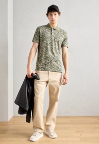 Grün gemustertes Polohemd mit kurzen Ärmeln, kombiniert mit beigen Hosen und weißen Sneakers. Eine schwarze Lederjacke liegt über einem Arm.