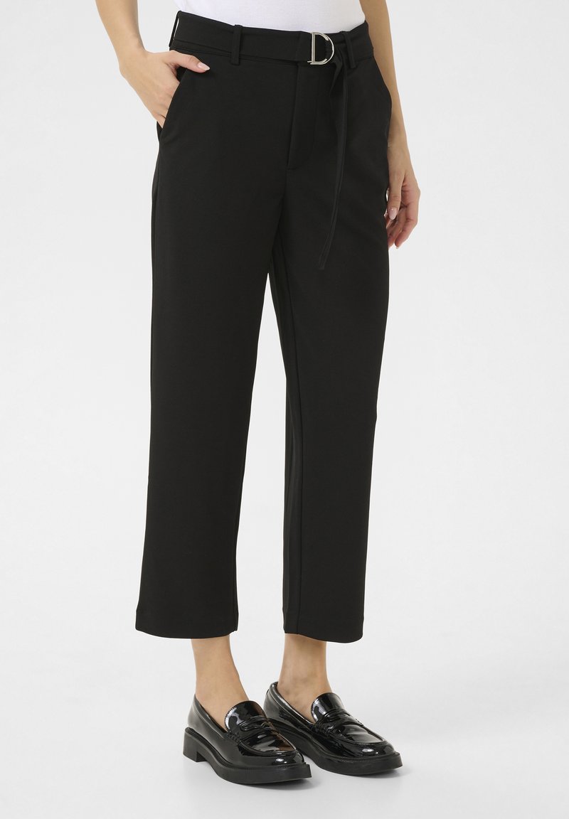 Zwarte taps toelopende broek van gladde stof, voorzien van een riem met een glanzende zilveren gesp en zijzakken. Zwart instap-schoenen met platte hak.