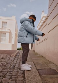 Niño con chaqueta acolchada azul claro, pantalones marrones y zapatillas blancas subiendo desde una calle adoquinada a la acera en un entorno urbano.