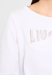 Vit skjorta med rund halsringning och ordet "LIU" i vita spetsbokstäver omgivna av små silvernitar.