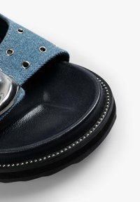 Denim schuifsandaal met een blauwe band met zilveren studs, een glanzend metallic accent en een zwarte gestructureerde zool met kralen details.