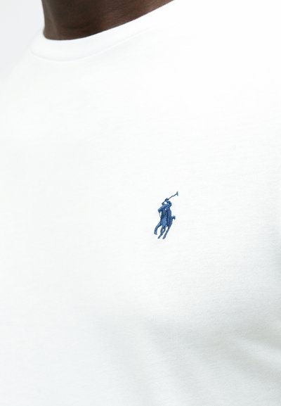 Biała bawełniana koszulka z niebieskim haftowanym logo gracza polo na lewej piersi. Okrągły dekolt, gładka tekstura, krótkie rękawy.