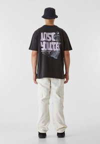 T-shirt noire avec un graphique audacieux "LOST YOUTH" en violet, portée avec un pantalon cargo crème et des chaussures noires, vue de dos.