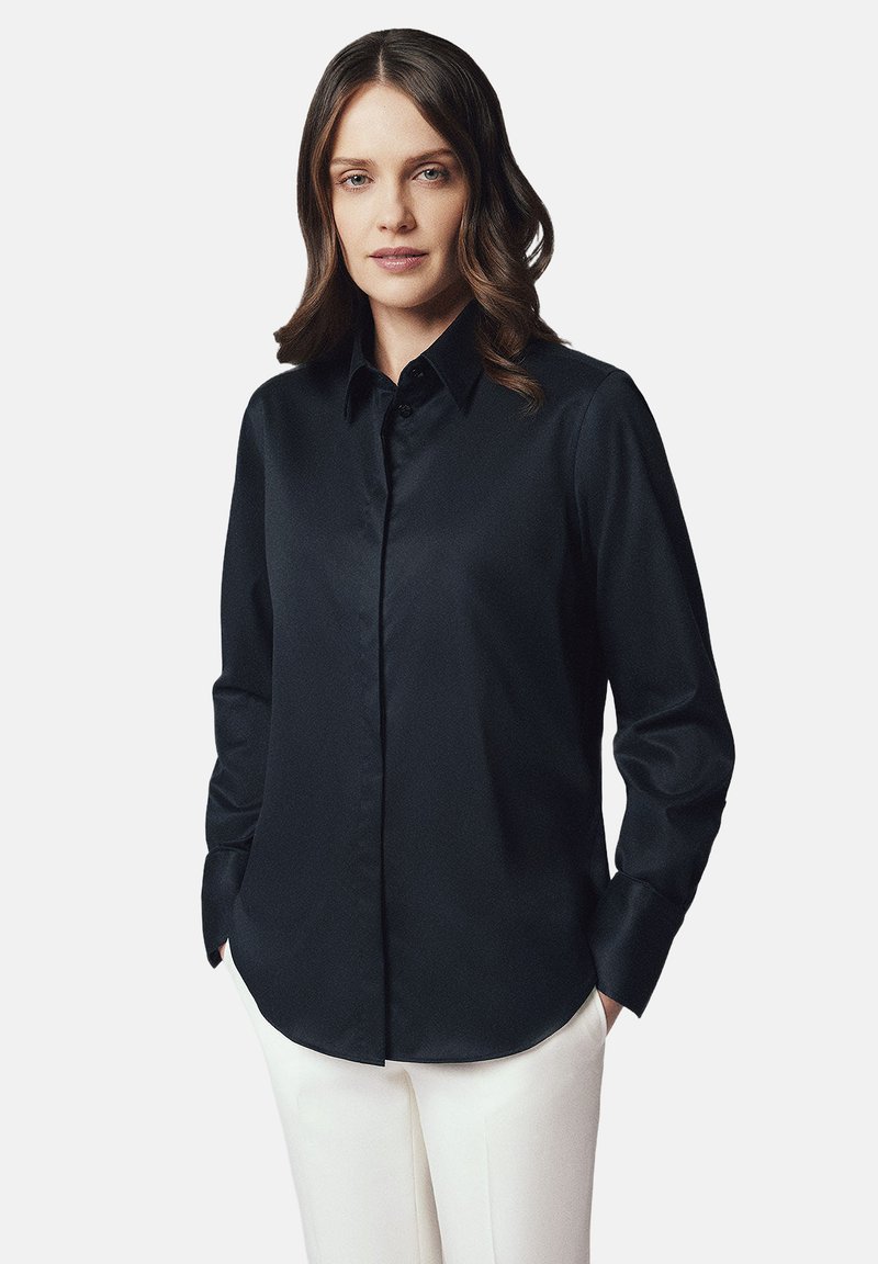 Czarna, długi rękaw koszula z gładkiego materiału, z kołnierzykiem, zapięciem na guziki i zaokrąglonym dołem. Zestawiona z białymi spodniami.