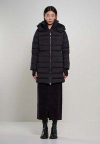Zwarte pufferjas met hoge kraag, ritssluiting en lange mouwen. Gekleed over een lange donkere rok en stevige zwarte laarzen. Matte textuur.