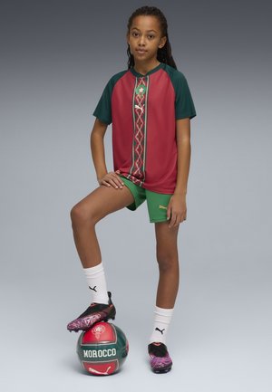Joven jugador de fútbol con el uniforme del equipo de Marruecos, apoyando un pie sobre un balón rojo y verde de Marruecos, frente a un fondo gris.