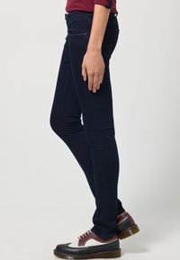 Jeans ajustados de mezclilla azul oscuro con una textura suave, con bolsillos y un ajuste de altura media, combinados con zapatos brogue bicolor.