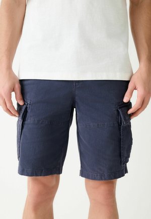 Muž nosiace námornícky modré cargo šortky s klopovými vreckami a biele tričko, stojaci pred jednoliatym bielym pozadím.