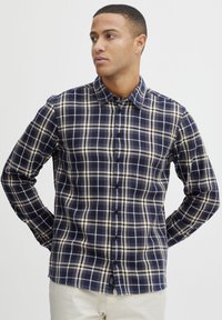 Cămașă cu mâneci lungi în carouri navy și crem, cu un guler clasic și o croială slim; prezintă nasturi în față și un tiv curbat.