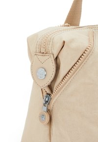 Angolo di una borsa di tessuto beige con cerniera, linguetta in pelle, bottone in metallo e manico in tessuto, su sfondo bianco.