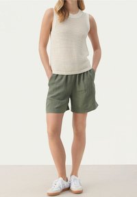 Beżowy bezrękawnik z dzianiny o otwartym wzorze, w połączeniu z zielonymi szortami i białymi trampkami z brązowymi podeszwami.