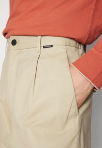 Pantaloni plissettati beige con un bottone nero e logo Calvin Klein. Indossati con una camicia a maniche lunghe arancione con polsini a contrasto.