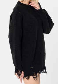Femme portant un pull en tricot noir oversize avec des bords et des poignets intentionnellement usés, debout devant un fond blanc uni.
