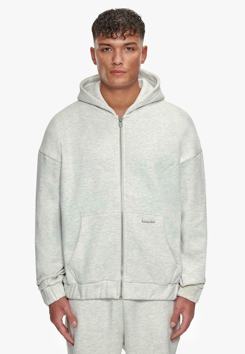 Haut à glissière gris fabriqué en tissu doux, doté d'une poche avant, d'une capuche et de poignets côtelés. Design minimaliste avec un petit logo en accent.