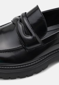 Mocassin en cuir noir poli avec semelle épaisse à relief et sangle décorative cousue sur le dessus.