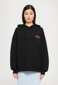 Mulher a usar um hoodie oversized preto com bolso frontal e o logótipo vermelho "FIORUCCI" no peito, combinado com uma saia azul clara.