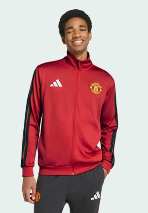 Smilende ung mand iført en rød Manchester United træningsjakke med sorte striber og matchende sorte sportbukser.