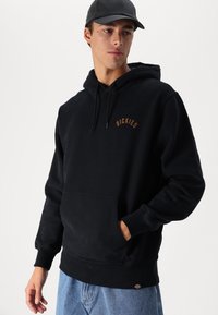 Sudadera negra con capucha, bolsillo frontal, puños y dobladillo acanalados. Cuenta con un logo beige en el pecho. Fabricada con un material de algodón suave y texturizado.