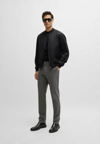 Svart bomberjacka över en svart skjorta, grå byxor och svarta ankelskor. Modellen bär mörka solglasögon. Släta, moderna material och design.