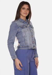 Giacca di jeans azzurra con design cropped, chiusura frontale con bottoni, due tasche sul petto e cuciture decorative. Texture liscia in tutto il capo.