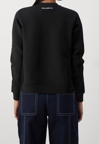 Sudadera negra de tela suave, con cuello redondo, logo blanco en la parte trasera, combinada con pantalones azul marino con costuras visibles y bolsillos.