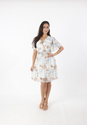 Kobieta stojąca, ubrana w białą, kwiatową sukienkę sięgającą do kolan z falbanowymi rękawami i beżowe szpilki z przezroczystymi paskami na białym tle.