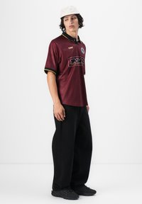 Jonge man met witte pet, bordeaux polo met korte mouwen en logo's, zwarte wijde broek en zwarte sneakers, staand tegen een effen witte achtergrond.